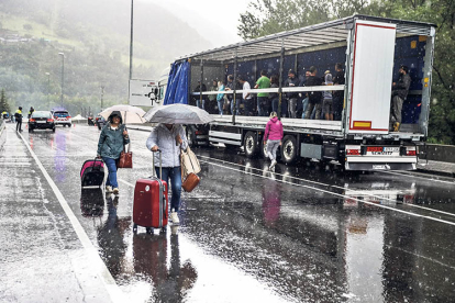 Turistes amb maletes creuant el tall de l’N-145 i manifestants resguardant-se de la pluja dins d’un camió.