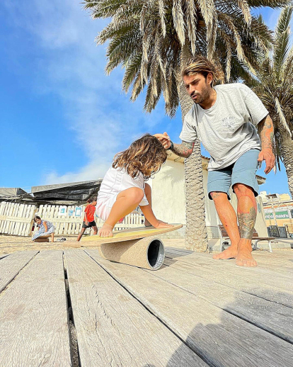 L'Albert Hinchado ensenya la seva filla, a la platja de Badalona, un matí d'estiu, a posar en pràctica amb una taula de fusta els moviments a fer per poder practicar surf i en un futur, potser, trepitjar un surf camp.